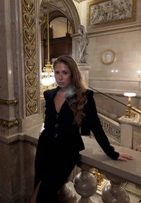 Czarny dopasowany strój ze złotymi guzikami, stojący przy marmurowej balustradzie. W tle ozdobna złoto-biała architektura.