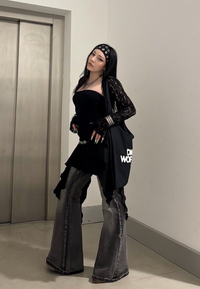 Mujer con top negro sin tirantes, mangas de encaje, vaqueros acampanados, diadema y pulseras posa cerca de las puertas cerradas de un ascensor llevando un bolso tote negro.