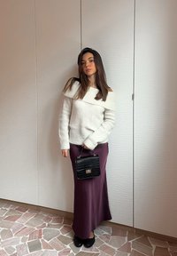 Maglione bianco a spalle scoperte, gonna maxi in satin viola, borsa strutturata nera, scarpe nere, in piedi contro uno sfondo bianco testurizzato.