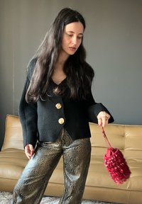 Cardigã preto de lã com botões dourados, combinado com calças brilhantes de padrão de cobra. Segurando uma bolsa vermelha com franjas, sentada num sofá beje.
