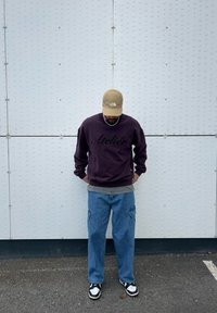 Sweat-shirt violet avec le texte "Atelier", sous-vêtement gris, pantalon cargo bleu et baskets noir et blanc. En arrière-plan, un mur blanc texturé.