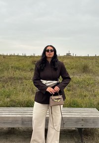 Hnědá saková bunda s bílým páskem, krémové kalhoty s širokými nohavicemi a malá hnědá kabelka. Tmavé sluneční brýle zvýrazňují kontrast outfitu.