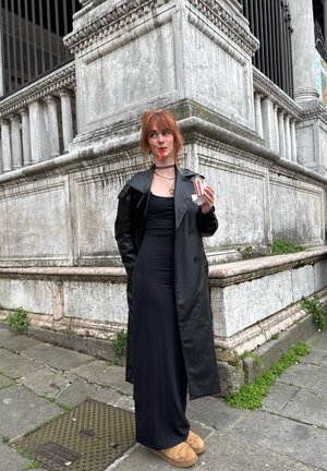 Joven con vestido negro y abrigo largo de cuero, de pie sobre un pavimento de piedra, sosteniendo palomitas y llevando zapatillas marrones cerca de la esquina de un edificio de piedra.
