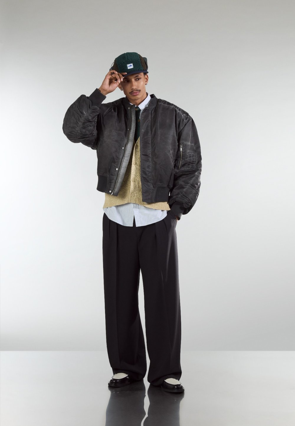 Black bomber jacket with quilted texture, layered over a beige jumper and white striped shirt. Loose black trousers and black shoes with white accents.