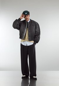 Black bomber jacket with quilted texture, layered over a beige jumper and white striped shirt. Loose black trousers and black shoes with white accents.