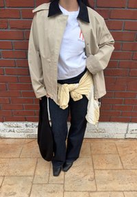 Personne portant une veste beige par-dessus un t-shirt blanc, un jean sombre, une veste claire nouée à la taille, tenant un sac noir, debout devant un mur de briques rouges.