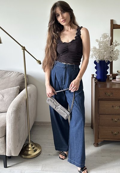 Top de tirantes negro con fruncidos, pantalones de mezclilla azul de pierna ancha, bolso gris con herrajes y sandalias planas negras, en un ambiente interior suavemente iluminado.