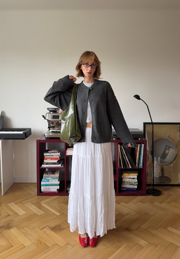 Persona con cabello corto y gafas que lleva un cárdigan gris oscuro, falda blanca, zapatos rojos y lleva una bolsa verde en una habitación con estanterías y lámpara.