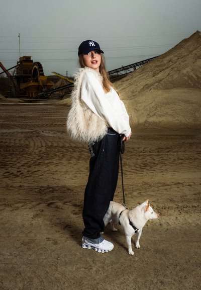 Chaleco de piel blanca sobre una sudadera blanca, jeans oscuros y holgados, y zapatillas blancas. Un pequeño perro blanco con correa está de pie al lado.