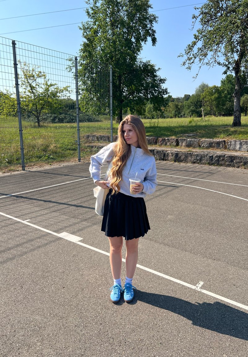 Jeune femme aux longs cheveux portant un sweat-shirt gris, une jupe noire et des baskets bleues, debout sur un terrain de basket en plein air et ensoleillé tenant une boisson.