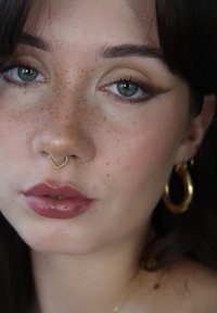 Nærbilde av et ansikt med lys hud og fregner, iført en gull septumring formet som et hjerte og store gull hoops. Leppene er glossede i rosa.