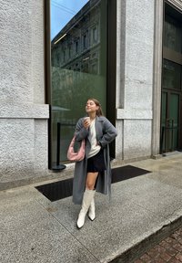 Grauer langer Mantel über einem hellen Pullover und einem schwarzen Rock, kombiniert mit weißen Knielangen Stiefeln und einer rosa Schultertasche. Kaffeebecher in der Hand.