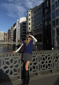 Colete de malha azul sobre uma camisa de mangas longas brancas, saia plissada cinza e botas pretas até ao joelho. Cenário urbano com edifícios e água.