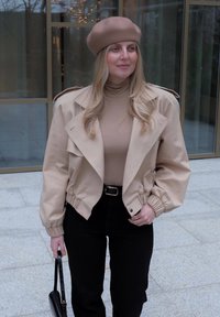 Veste courte beige avec manches bouffantes, col montant et détails boutonnés, associée à un pull à col roulé beige et un pantalon taille haute noir.