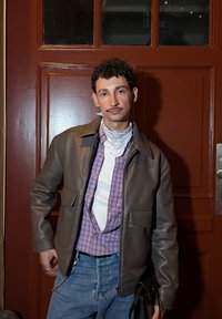 Ung man med lockigt hår och mustasch som bär en brun läderjacka, lila rutiga skjorta, vit bandana och jeans står framför en röd dörr.