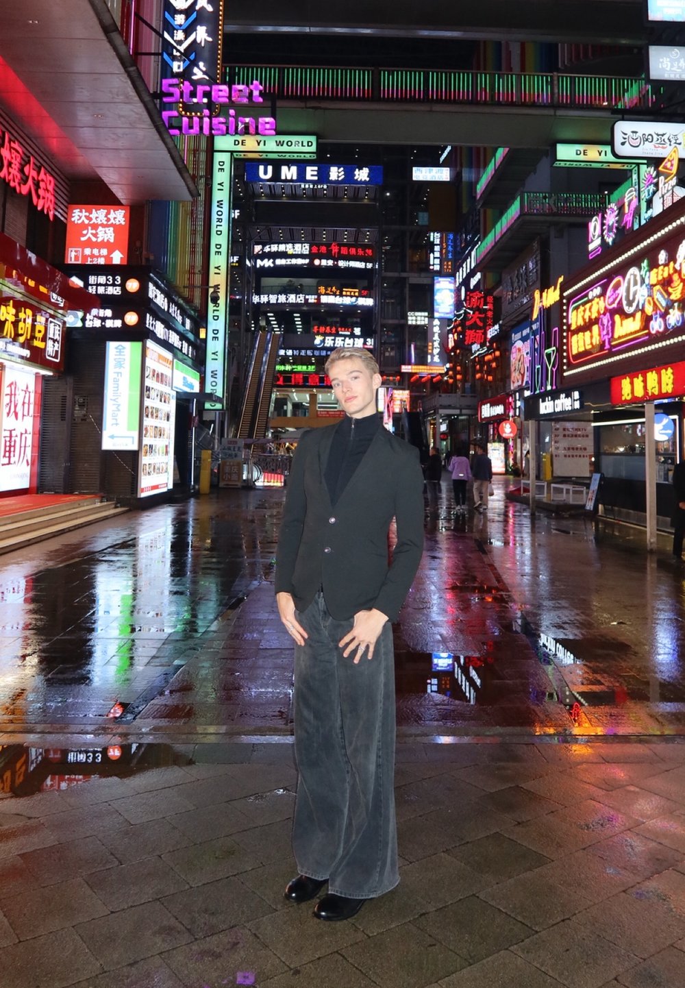 Una persona è in piedi su una strada bagnata, indossa un blazer nero e pantaloni grigi a gamba larga, circondata da insegne al neon illuminate vivacemente.