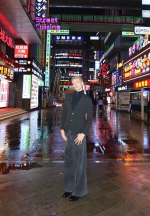Una persona está de pie en una calle mojada, vestida con una chaqueta negra y pantalones gris de pierna ancha, rodeada de señales de neón muy iluminadas.