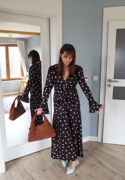 Mujer con vestido blanco con lunares negros que sostiene un bolso marrón, de pie cerca de un espejo de cuerpo entero en una habitación con paredes azul claro.