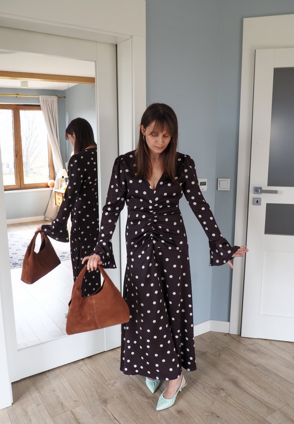 Femeie în rochie alb-negru cu buline, ținând o geantă maro, stând lângă o oglindă în toată înălțimea, într-o cameră cu pereți albastru deschis.