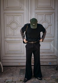 T-shirt noire surdimensionnée portée par-dessus une chemise gris à manches longues, associée à un pantalon noir à jambes larges et des chaussures marron. Casquette verte et sac à bandoulière.