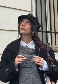 Jeune femme aux cheveux tressés portant une casquette noire, un pull gris par-dessus une chemise rayée, tenant des lunettes de soleil, debout devant la façade d'un bâtiment.