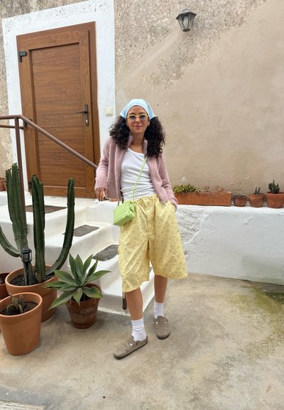 Cárdigan fuzzy rosa claro, camiseta blanca de tirantes, pantalones cortos amarillos con estampado, bolso cruzado verde, calcetines blancos, zapatos marrones, de pie junto a plantas en macetas.