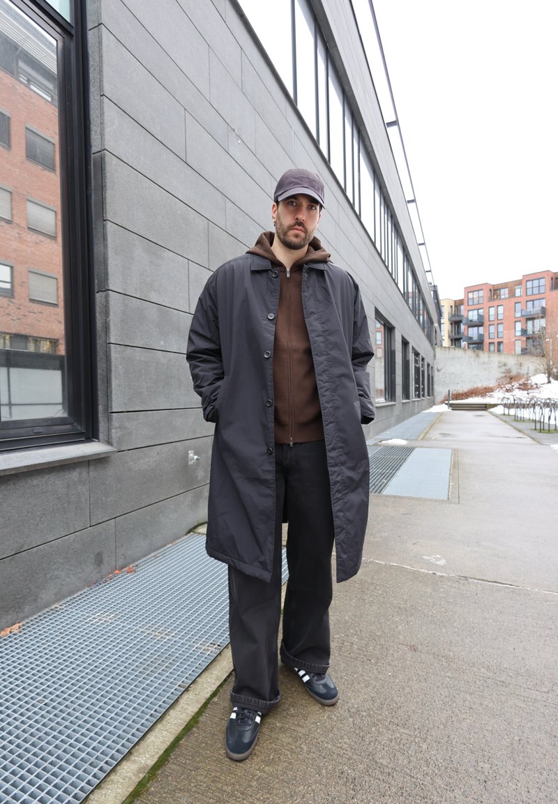 Mand iført en sort frakke, brun hættetrøje, sorte bukser og kasket, der står på en belagt sti ved siden af en moderne grå bygning.