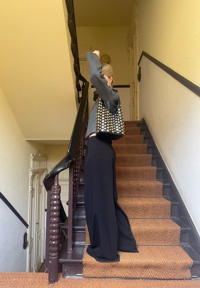 Pantalones negros de pierna ancha con una blusa gris, llevando un bolso de cuero con tachuelas doradas, de pie en una escalera marrón con patrón de espiga.