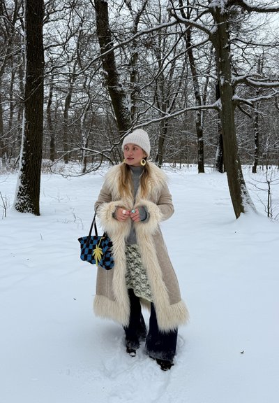 Mujer con abrigo crema con borde de piel, gorro de punto blanco y pantalones oscuros, sosteniendo una bolsa azul a cuadros, de pie en un bosque nevado con árboles desnudos.