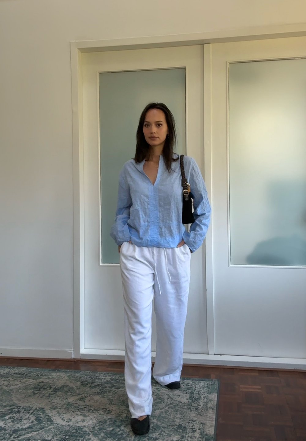 Lichtblauw, gestructureerd shirt met lange mouwen, witte broek met koordjes en zwarte schoenen. Staand voor deuren van gezandstraald glas op een houten vloer.