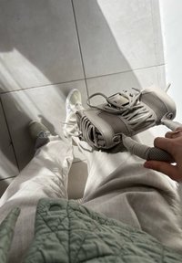 Beige Sneaker mit Mesh- und Gummidetails, gehalten von einer Hand. Eine graue gesteppte Jacke und helle Hosen sind im Vordergrund zu sehen. Kachelfußboden darunter.