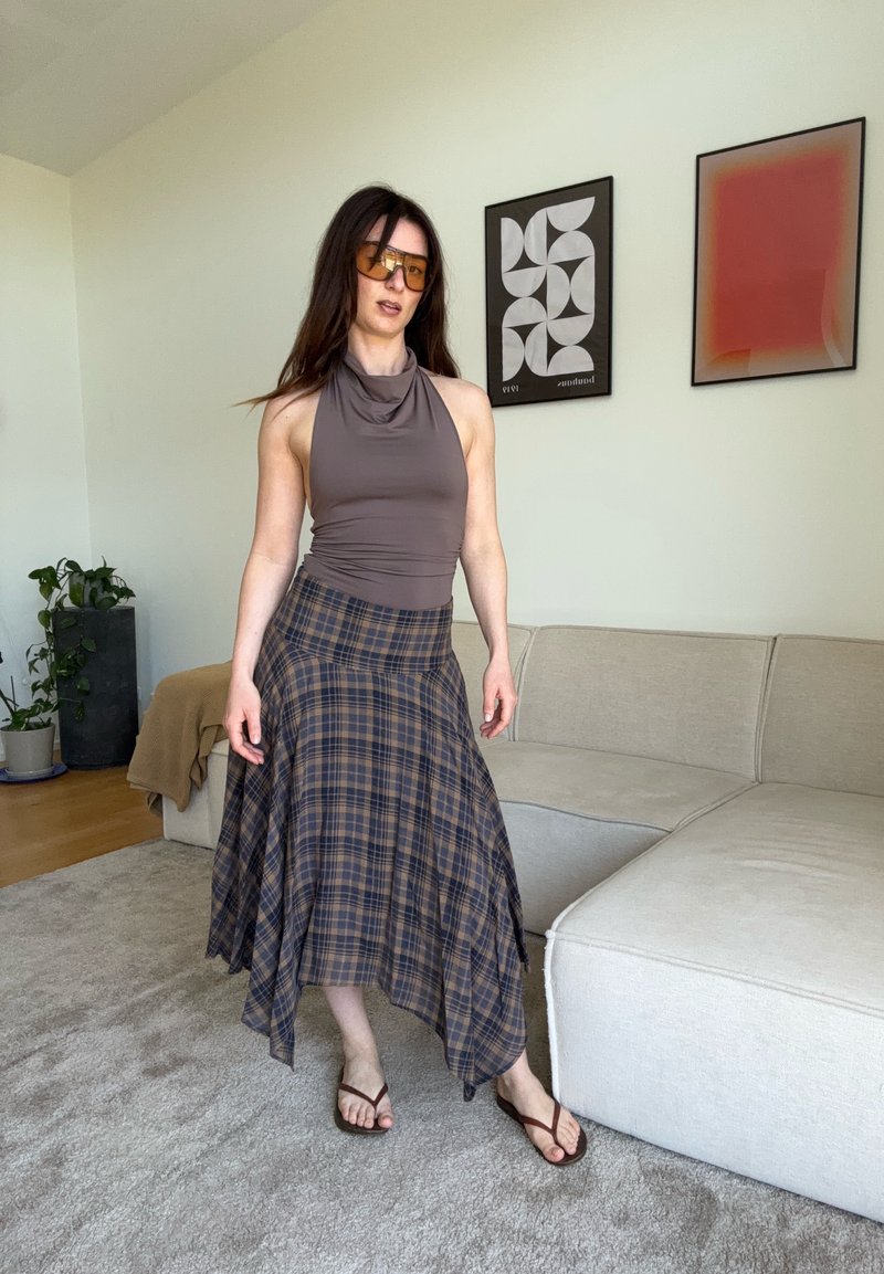 Frau steht im Wohnzimmer, trägt ein ärmelloses braunes Top, asymmetrischen karierten Rock, Flip-Flops und große getönte Sonnenbrillen.