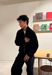 Νεαρό άτομο με σκούρα μαλλιά και τατουάζ στα χέρια, φορώντας μαύρο πουλόβερ, στέκεται κοντά σε ξύλινο τραπέζι με κούπα σε μινιμαλιστικό δωμάτιο.