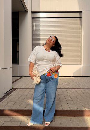 Mujer con cabello largo y oscuro, lleva una camiseta blanca, camisa colorida debajo, vaqueros de pierna ancha, zapatos blancos abiertos en los dedos, gafas de sol y sostiene un bolso beige al aire libre.