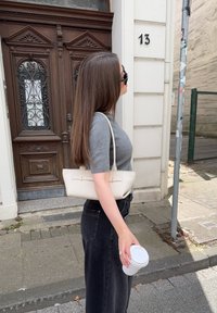 Una mujer con cabello largo y castaño lleva una blusa gris de manga corta y jeans negros, sosteniendo una taza blanca al lado de un pequeño bolso de crema.