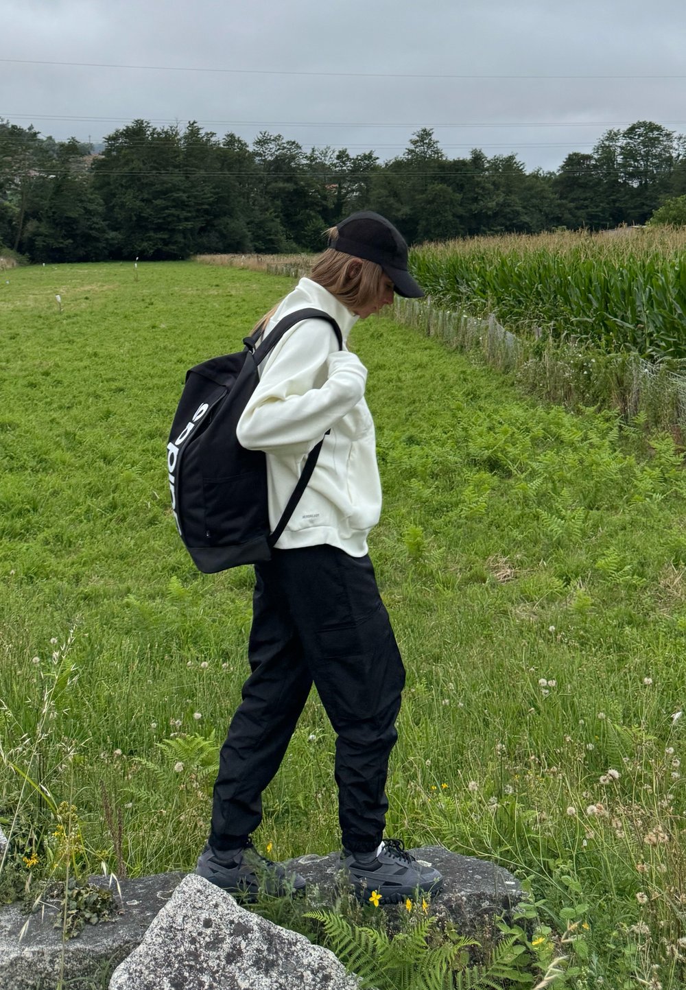 Zaino nero, giacca bianca con zip, pantaloni cargo neri; in piedi su una roccia con erba verde e un campo di mais sullo sfondo.