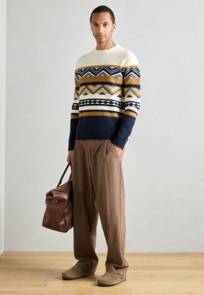 Suéter de punto con parte superior crema, secciones con patrón en azul marino y mostaza; pantalones marrones; sostiene un bolso de cuero marrón; textura suave.