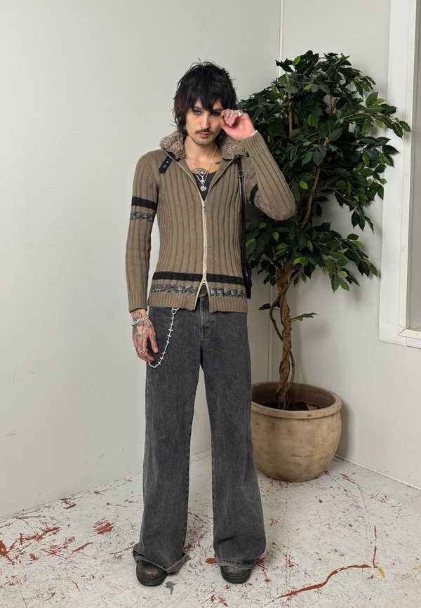 Homme aux cheveux noirs ébouriffés portant un pull à fermeture éclair rayé marron, un jean large gris et des bracelets, debout à côté d'une plante feuillue en pot.