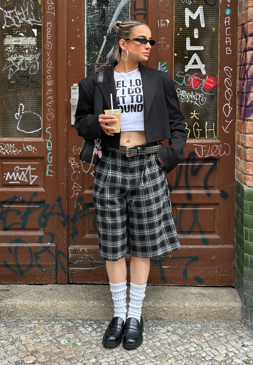 Sacou negru scurt, top scurt alb cu grafică, pantaloni scurți în carouri și loafers negri. Accesoriile includ cercei hoop și o băutură în mână.