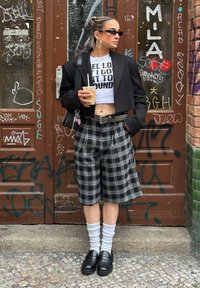 Sacou negru scurt, top scurt alb cu grafică, pantaloni scurți în carouri și loafers negri. Accesoriile includ cercei hoop și o băutură în mână.