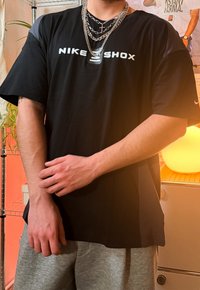 Svart Nike Shox-t-shirt med korta ärmar och en vit logga på framsidan. Bärs med metalliska lager på kedjor. Gråa mjukisbyxor syns.