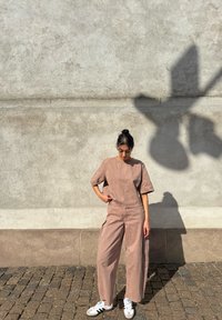 Ung kvinde i en løs brun dragt og hvide sneakers, som står på en brostensbelagt gade foran en struktureret beige væg i sollys.