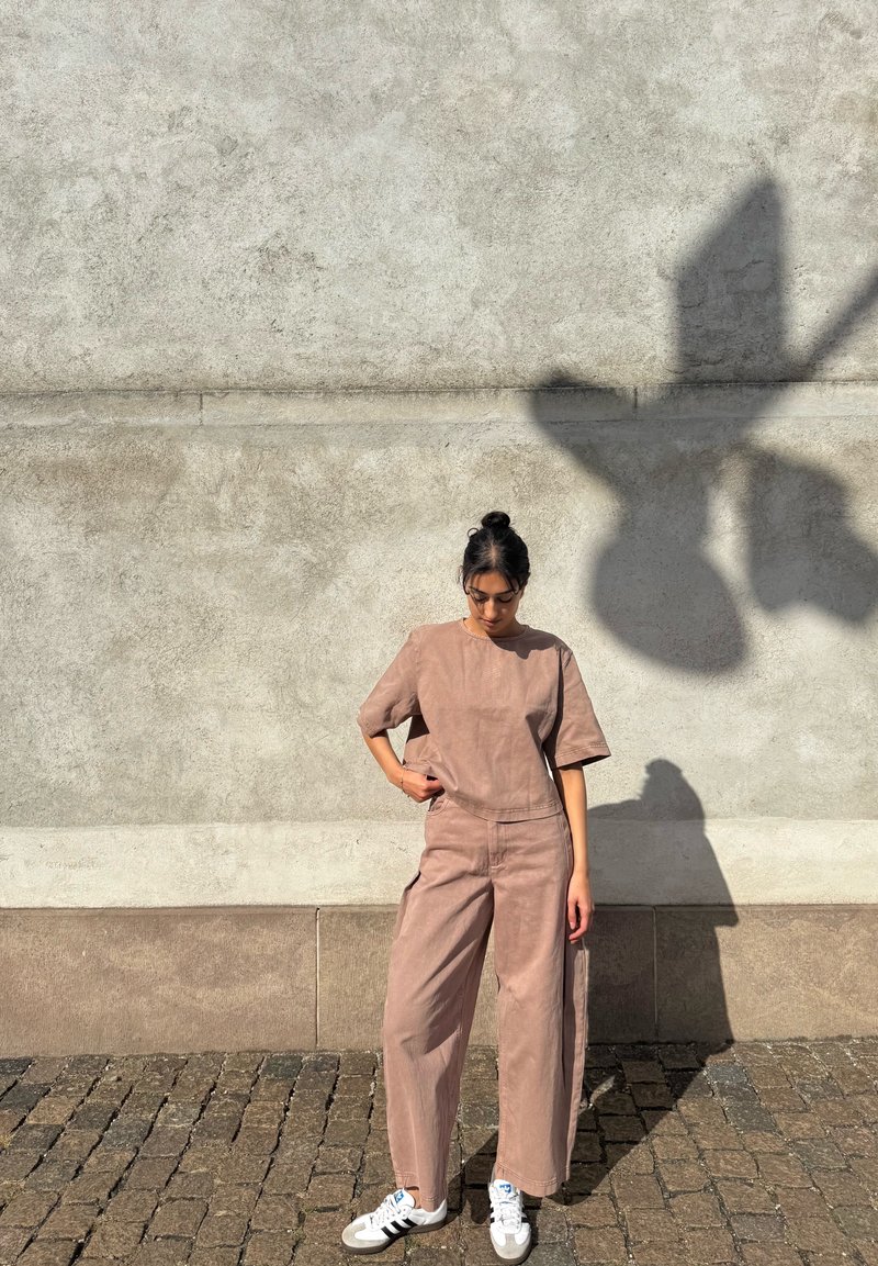 Young woman in a loose brown outfit and white sneakers, standing on a cobblestone street against a textured beige wall in the sunlight.