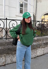 Sweat à capuche vert avec une poche kangourou, associé à un jean en denim bleu clair. Sac à bandoulière noir et casquette verte ornée d'un logo blanc. Environnement extérieur.
