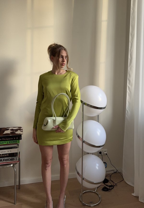 Femme en robe verte tenant un sac à main blanc, debout à côté d'une lampe sur pied moderne avec trois lumières sphériques dans une pièce minimaliste.