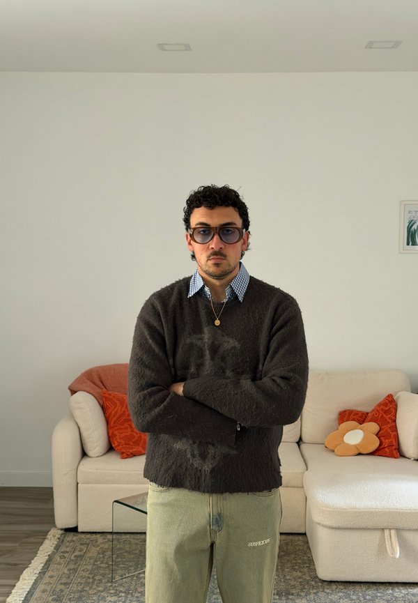 Uomo che indossa occhiali da sole, maglione scuro e pantaloni chiari, in piedi con le braccia incrociate in un soggiorno con un divano bianco e cuscini arancioni.