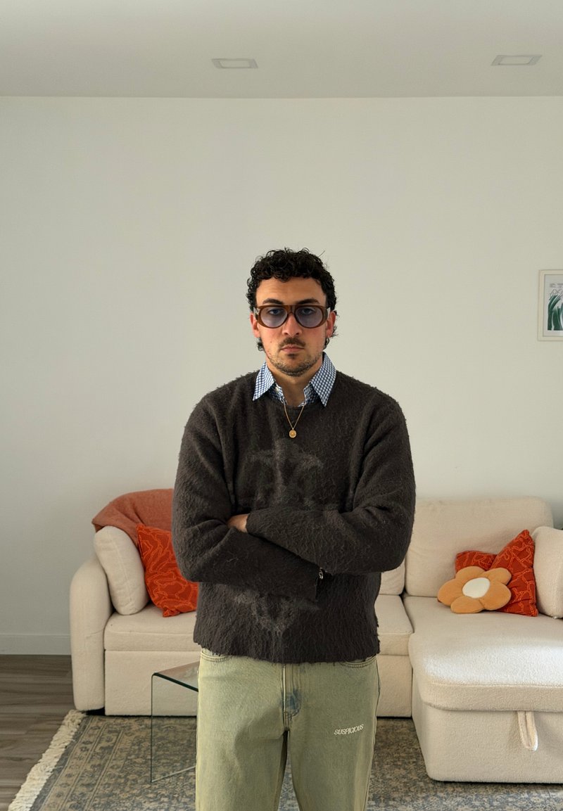 Homem a usar óculos de sol, suéter escuro e calças claras, está de pé com os braços cruzados numa sala de estar com um sofá branco e almofadas laranja.