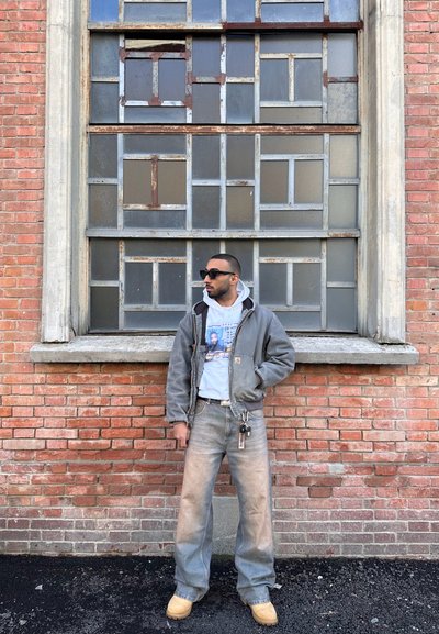 Hombre con gafas de sol, chaqueta gris, sudadera gráfica, jeans anchos y botas beige se encuentra frente a una pared de ladrillo con una gran ventana de múltiples paneletas.