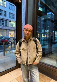 O ținută relaxată formată dintr-o jachetă beige din catifea cu coastă purtată peste o cămașă în carouri, combinată cu blugi albaștri deschis și o șapcă roz, stând în interior, lângă o fereastră.