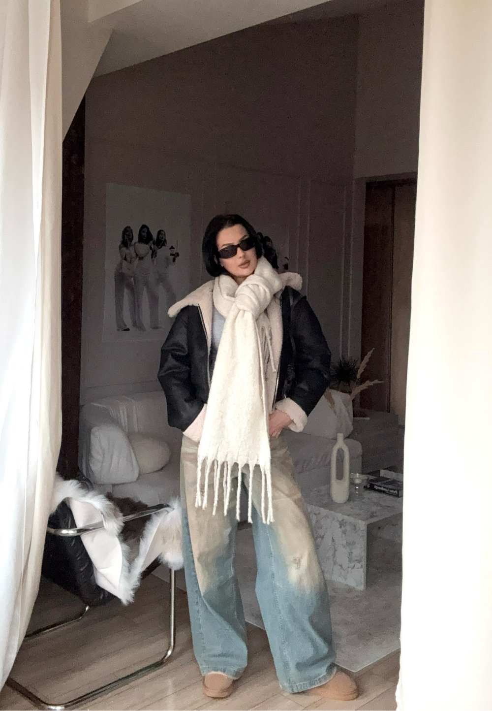 Jachetă neagră din piele ecologică cu guler de blană, eșarfă mare crem cu franjuri, blugi largi bleu deschis și pantofi slip-on bej.